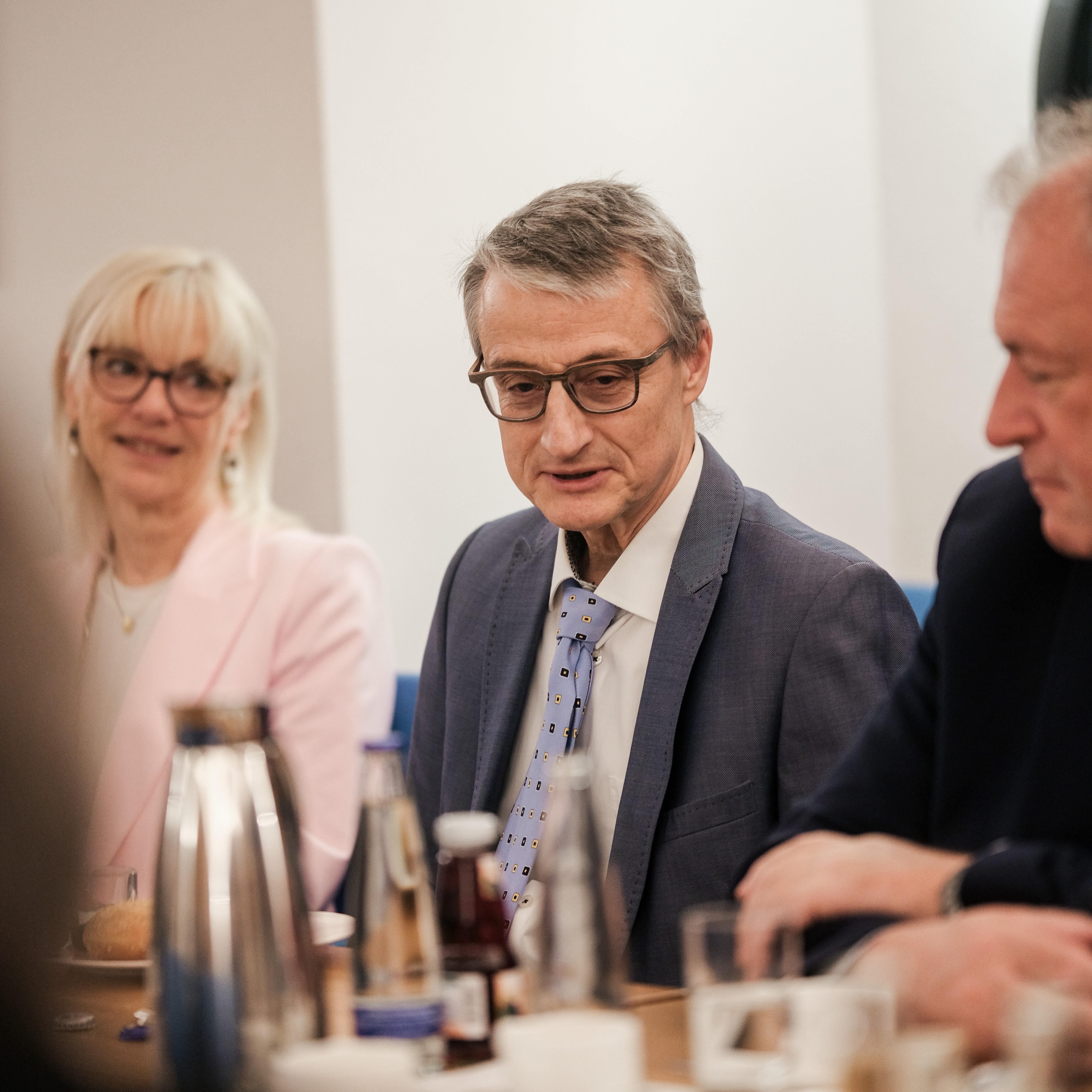 Ein Mann im Anzug sitzt an einem Besprechungstisch. Rechts und links von ihm sind verschwommen weitere Personen zu sehen.
